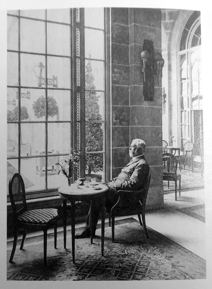 Black and white photograph of a man in a suit sitting at a small table beside a tall window in the Adelphi Hotel in 1912
