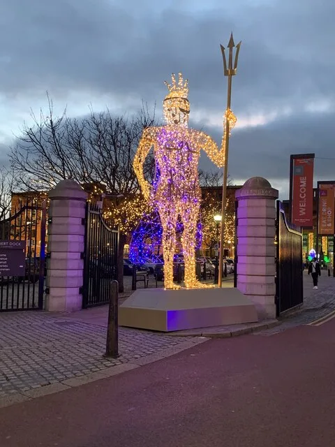 A tall illuminated figure holding a trident, made of warm white lights, standing at the gated entrance to the Royal Albert Dock at dusk