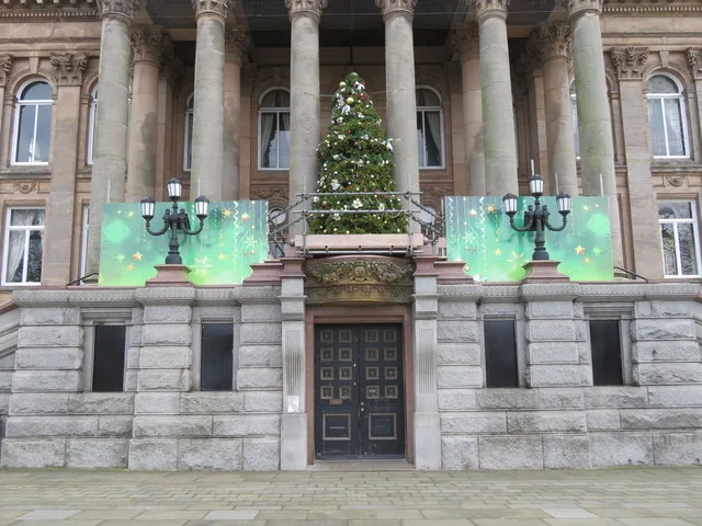 A decorated Christmas tree on the balcony above the entrance of Birkenhead Town Hall, between stone columns, with green lit panels either side