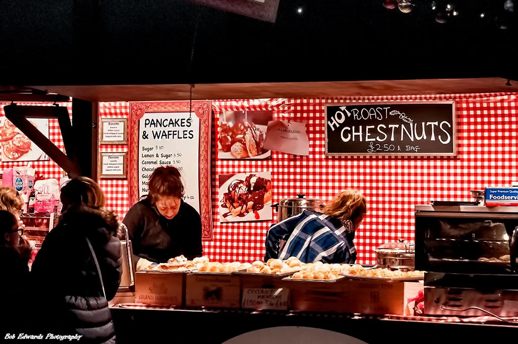 A Christmas market stall with red gingham walls selling hot roast chestnuts, pancakes and waffles, with customers queuing at the counter