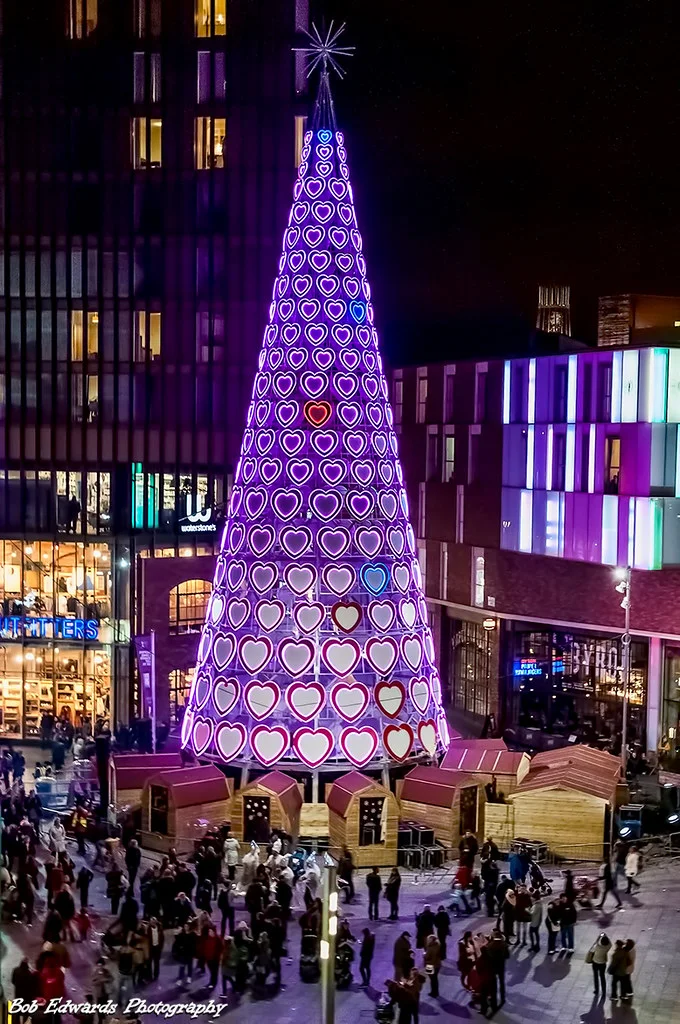 A tall cone-shaped Christmas tree covered in glowing pink and purple heart lights at Liverpool ONE at night, with crowds and market huts around its base
