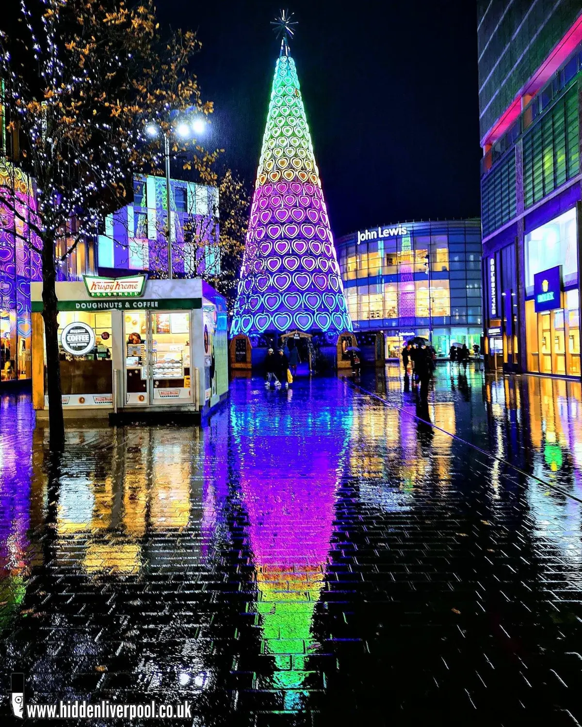 Liverpool ONE's heart-light Christmas tree glowing purple, pink and green on a rainy night, reflected in the wet paving, with John Lewis and a Krispy Kreme kiosk alongside