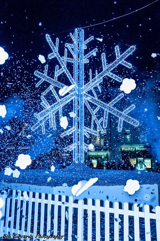 A giant illuminated snowflake sculpture glowing blue and white above a white picket fence in Williamson Square, with artificial snow in the air