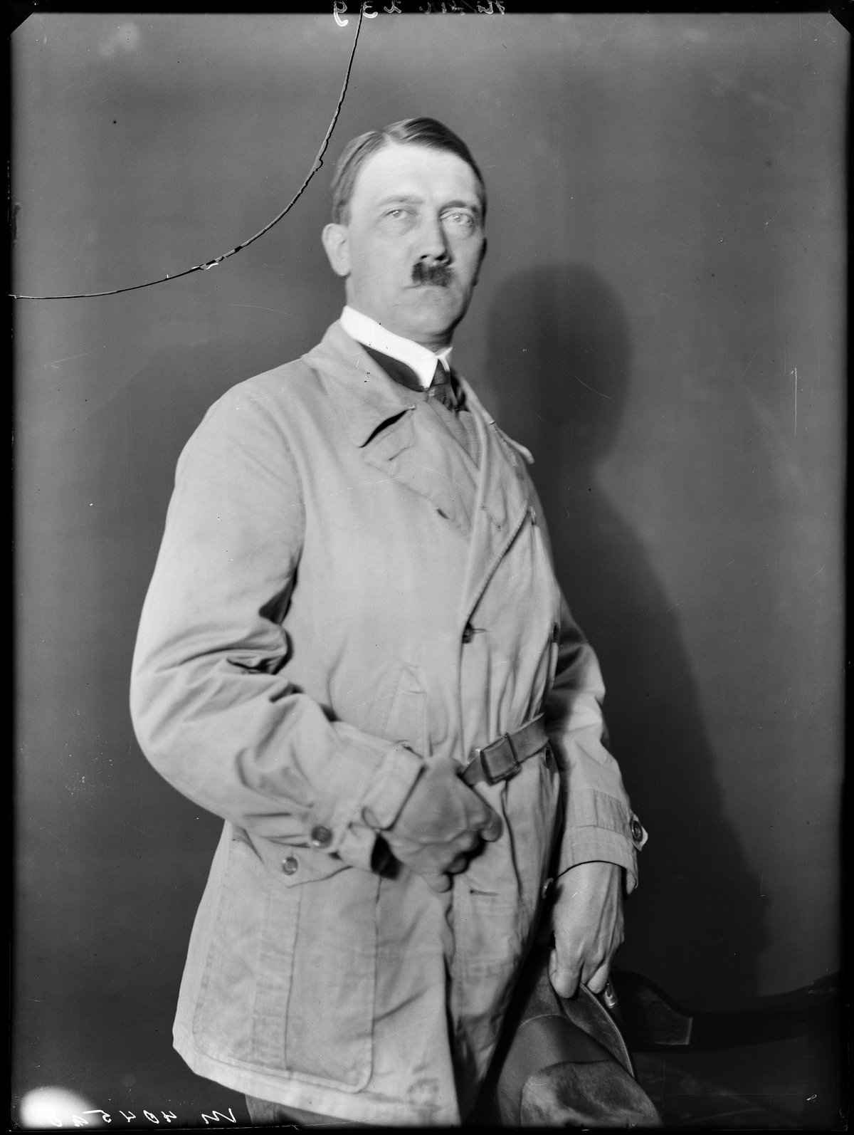 Black and white studio portrait of Adolf Hitler in the 1920s, wearing a coat and holding a hat.