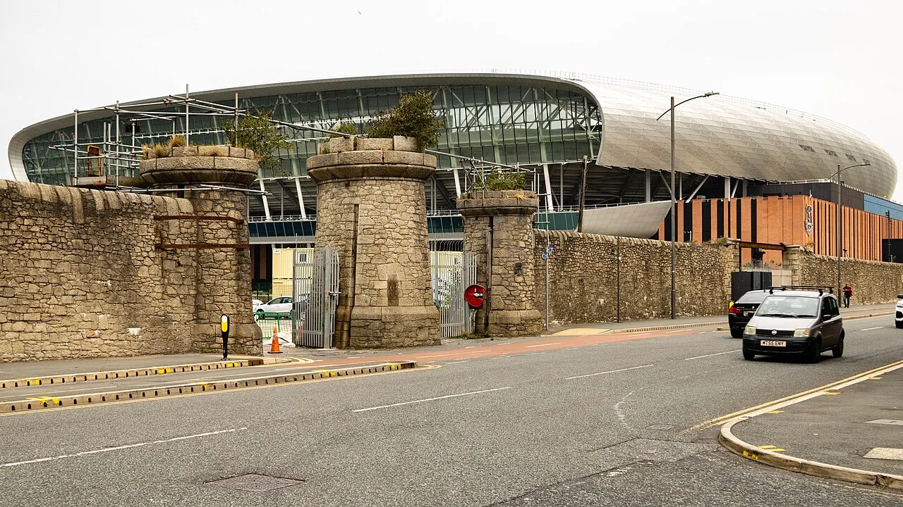 Stone gate pillars and a low stone wall beside Regent Road, with the stadium's glass and silver roof behind