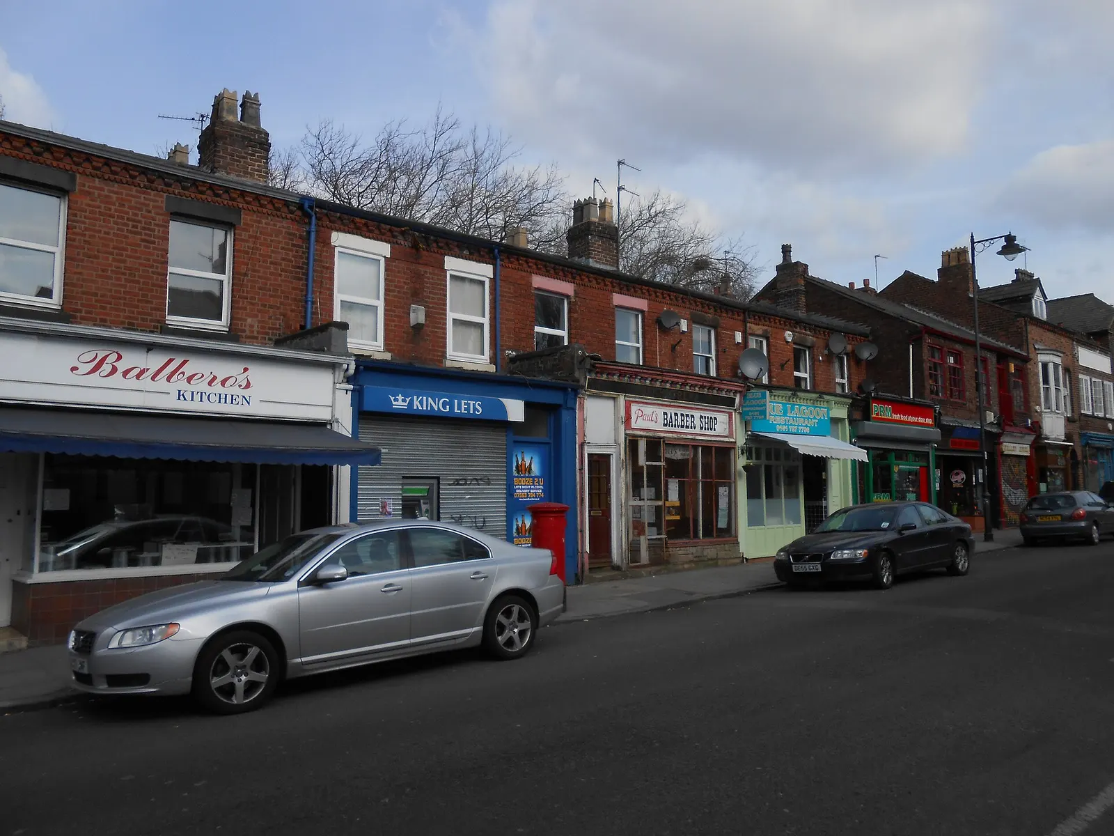 A row of shops and a barber on Lark Lane with cars parked along the kerb outside