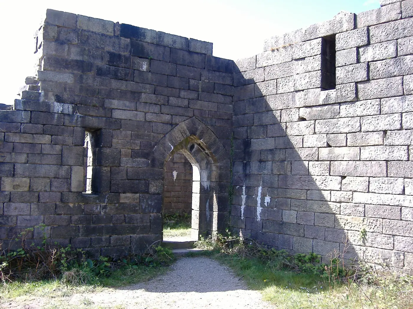 A ruined looking stone castle with round corner towers and empty window openings, standing on grass beside a reservoir