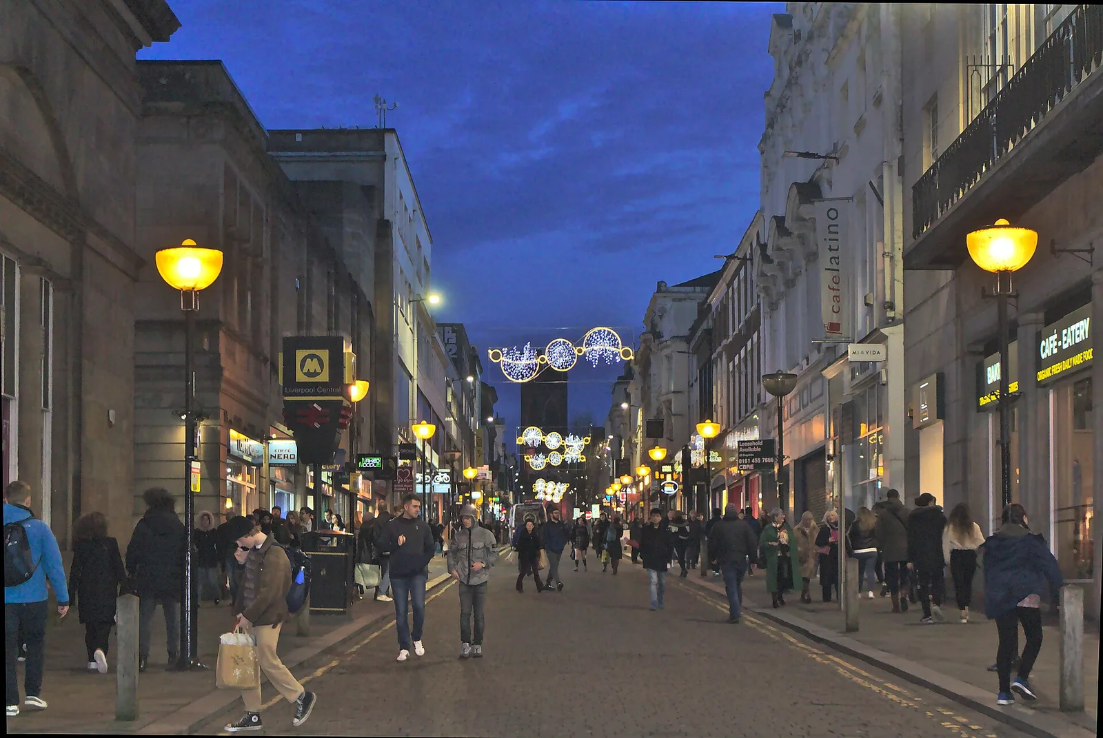 Shoppers walking up Bold Street in Liverpool at dusk beneath Christmas street lights, with the Liverpool Central station sign on the left