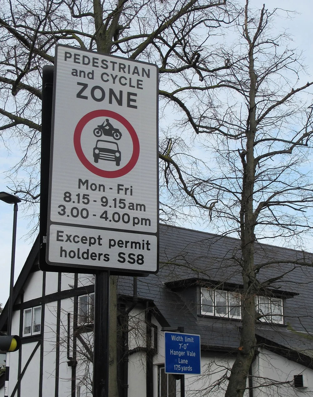 A School Street sign on a pole beside a residential road, reading Pedestrian and Cycle Zone, with a no-cars-or-motorcycles roundel, the hours Mon to Fri 8.15 to 9.15am and 3.00 to 4.00pm, and a plate reading Except permit holders