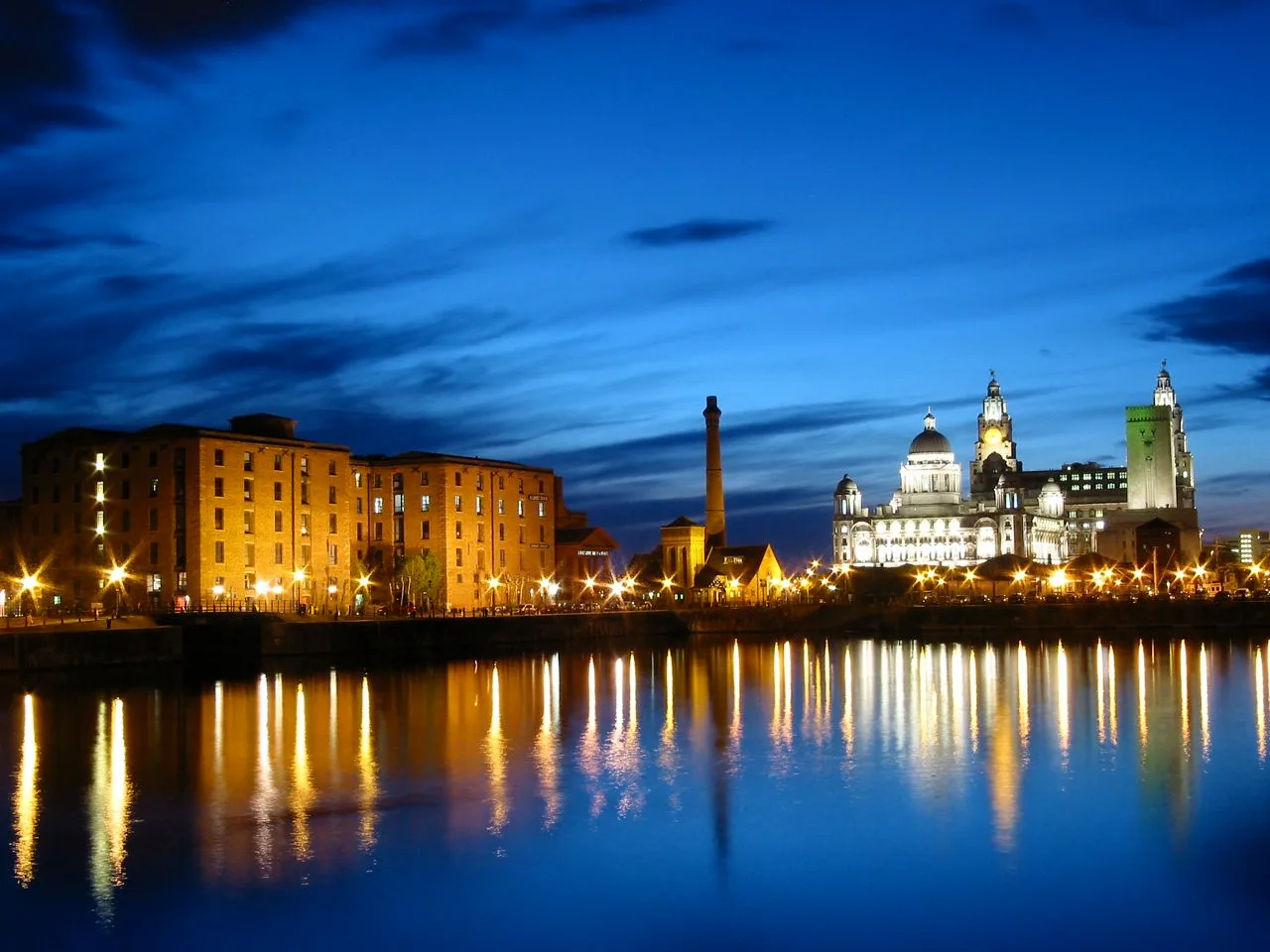 Royal Albert Dock warehouses and the Pumphouse lit at dusk, with the Port of Liverpool and Royal Liver buildings beyond and lights reflected in the dock water