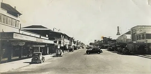 A wide street of canopied shopfronts with parked period cars