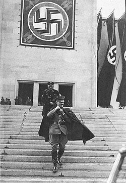 A uniformed figure walking down steps beneath large swastika banners
