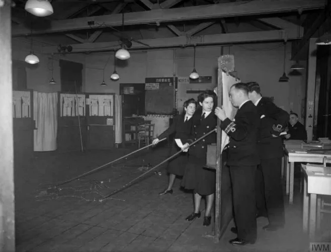 Wrens and naval officers holding long pointers in a large wartime training room