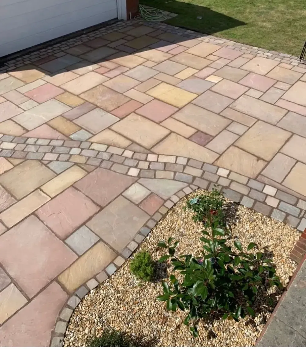 Multi-coloured sandstone paving with a cobble border curving round a gravel bed, beside a white garage door