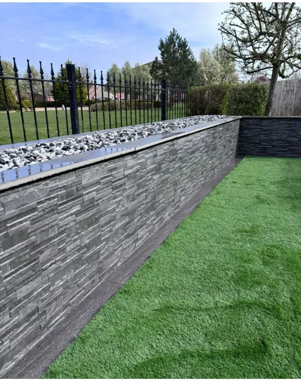 Grey stone-effect garden wall with a gravel-topped planter, beside a lawn of artificial grass and black railings
